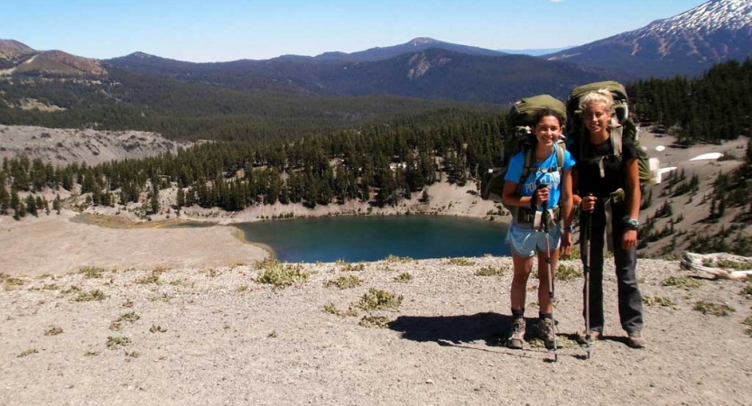 Two students wearing backpacks smile at the camera. There is a blue lake and mountains in the background. 